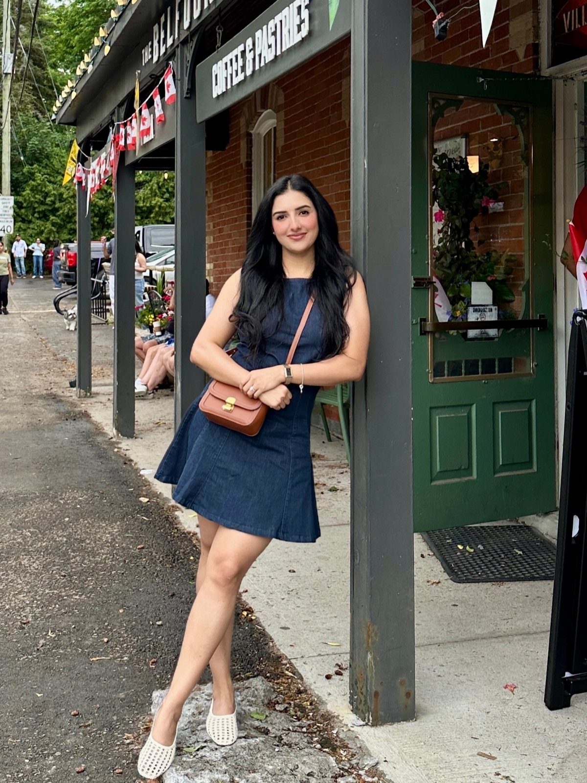 A young woman in a blue denim dress and white flats posing by a coffee shop.
