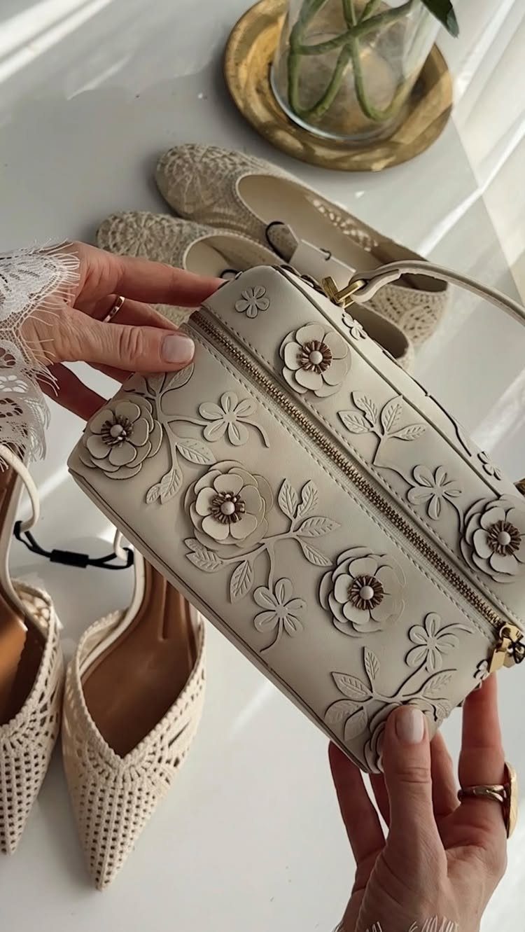 Hands holding a white floral embossed leather handbag near white patterned flats.