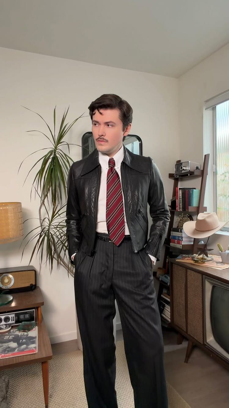 Man in a black leather jacket, red striped tie, and black high-waisted pinstripe trousers standing indoors.