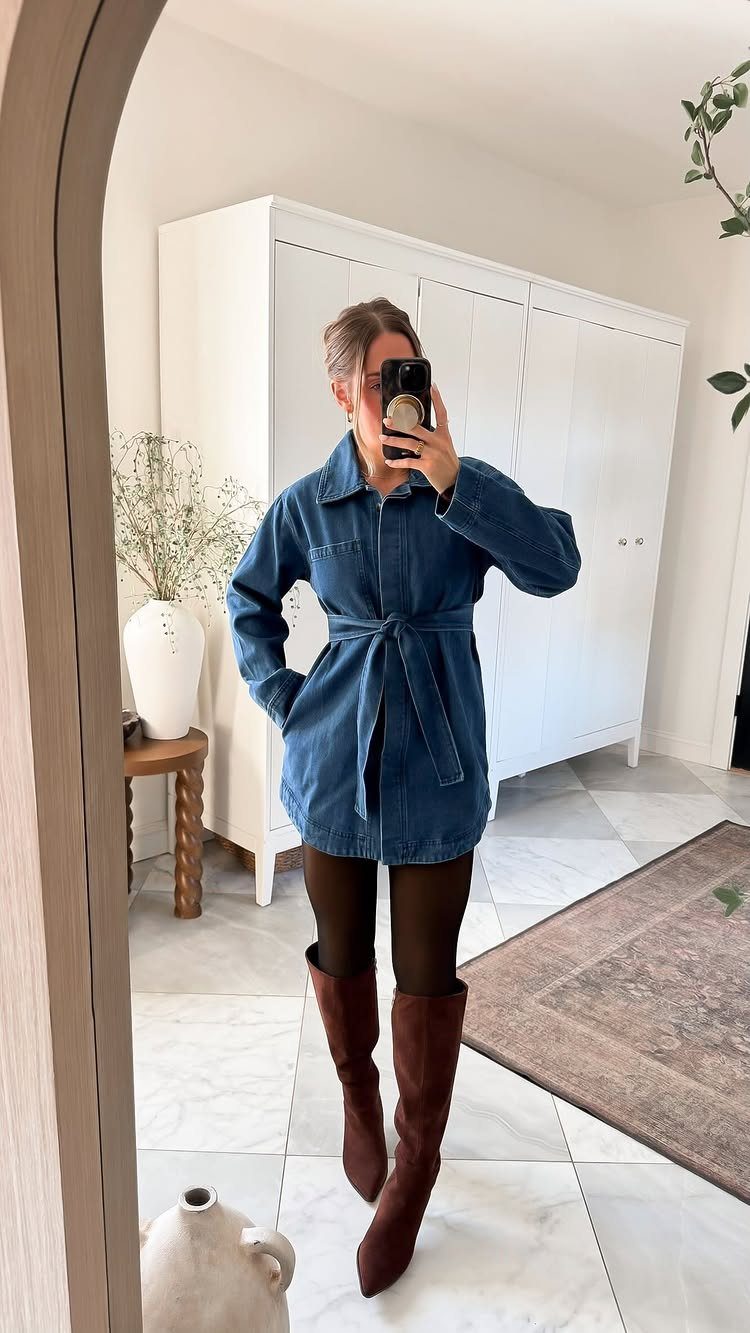 Woman in a denim shirt dress with brown boots posing in a mirror.