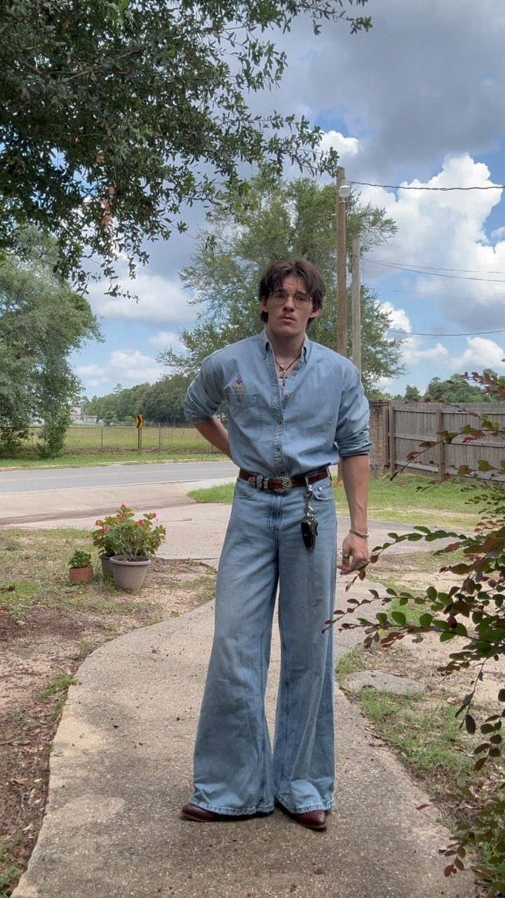 A man wearing a denim shirt and wide-leg denim jeans standing on a sidewalk outdoors.