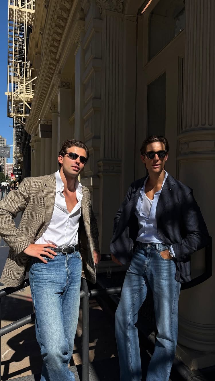 Two men posing in denim jeans, white shirts, and stylish tailored blazers outdoors.