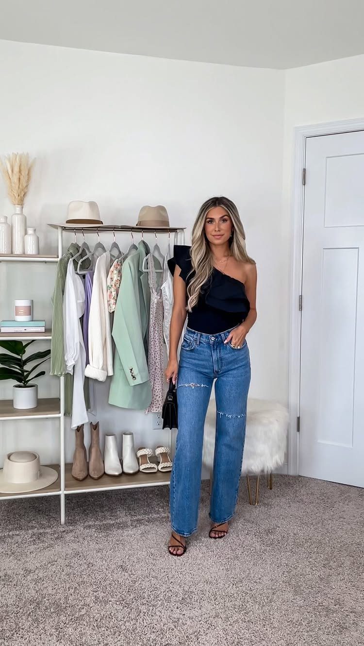 Woman wearing a black one-shoulder ruffled top and blue denim jeans standing in a room.