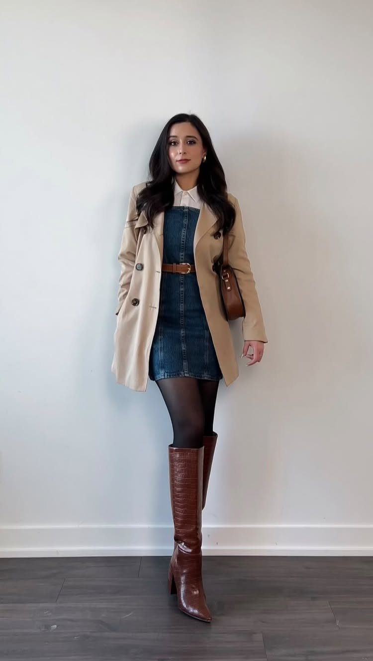 Woman posing in a denim dress, tan trench coat, and tall brown leather boots.