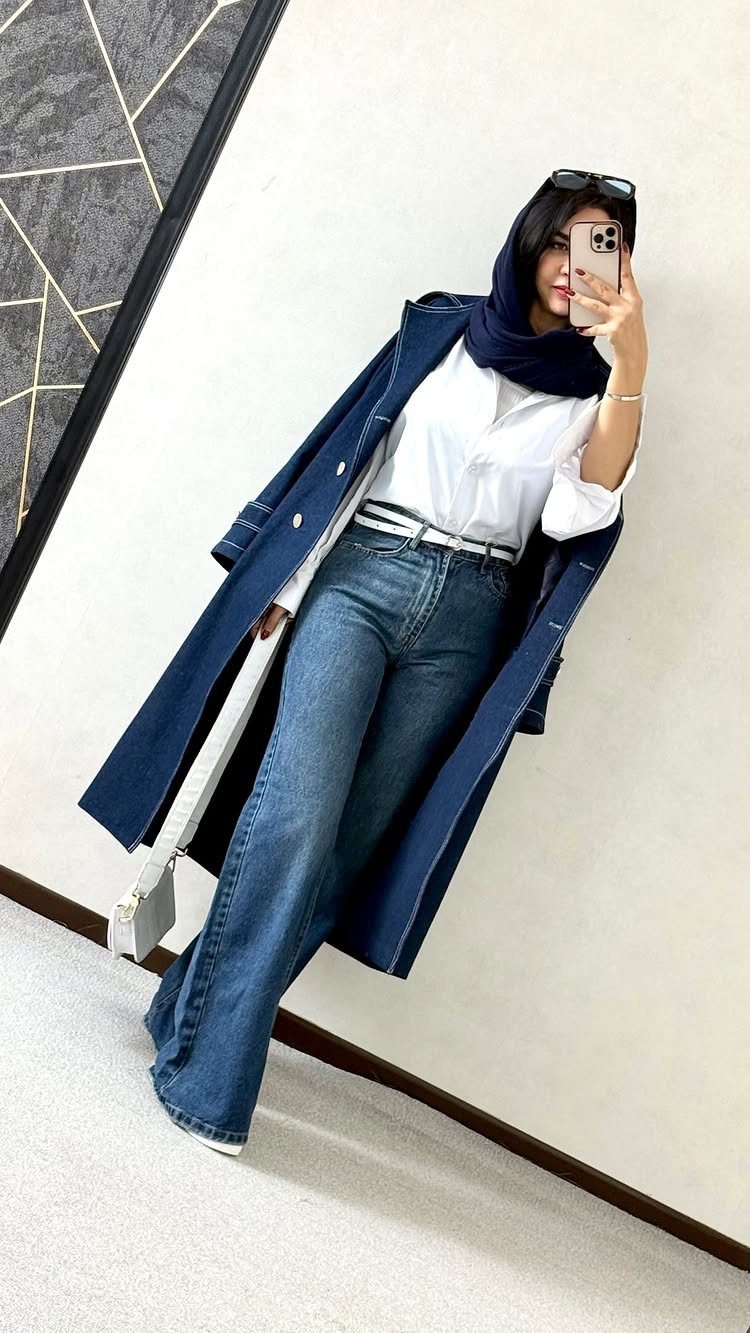 A woman posing in a mirror wearing a denim coat, white shirt, and flared denim jeans.