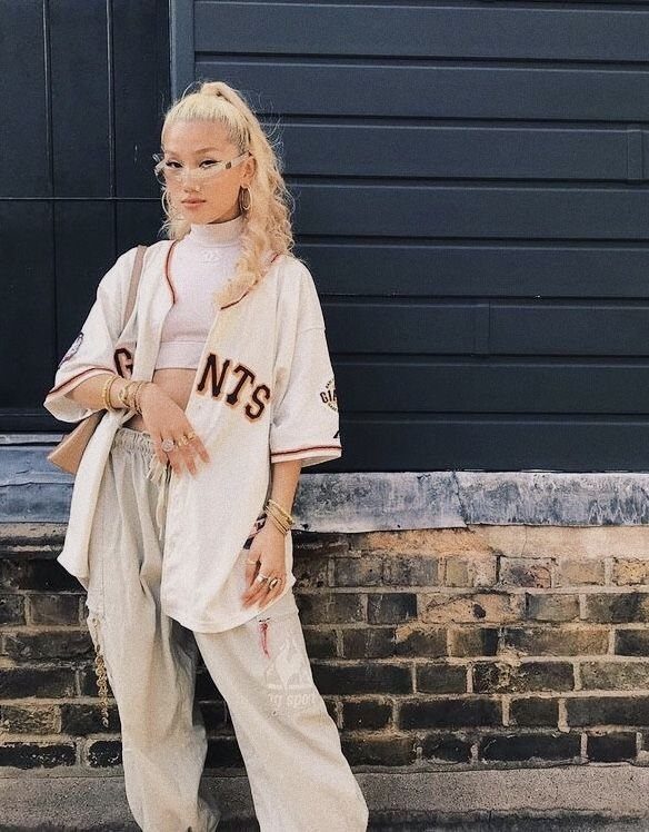 Woman in an oversized baseball jersey, pink crop top, and baggy pants posing outdoors.