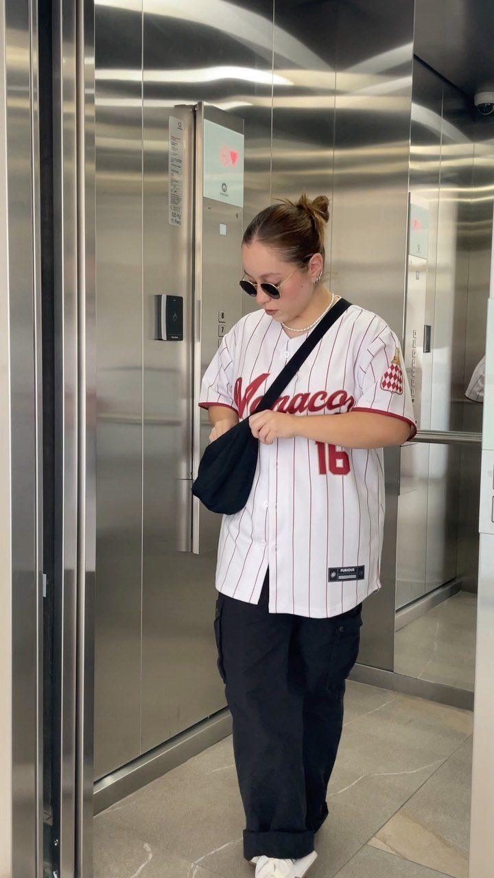 A woman wearing a white pinstriped baseball jersey and black cargo pants in an elevator.