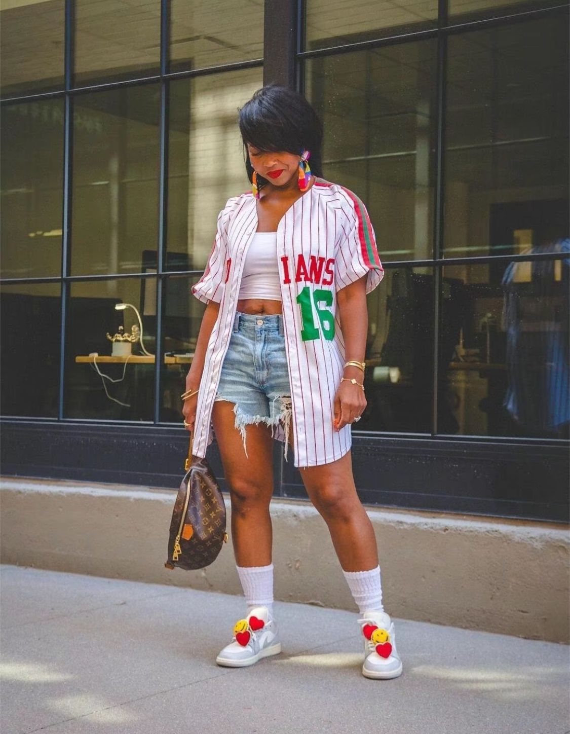 Woman wearing a pinstriped baseball jersey, denim shorts, and white sneakers standing on a sidewalk.