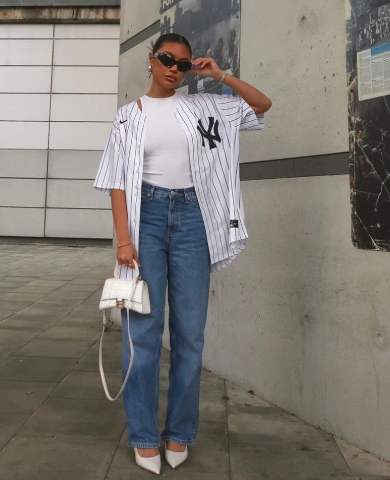 A woman posing in a pinstriped baseball jersey, blue jeans, and white heels holding a handbag.