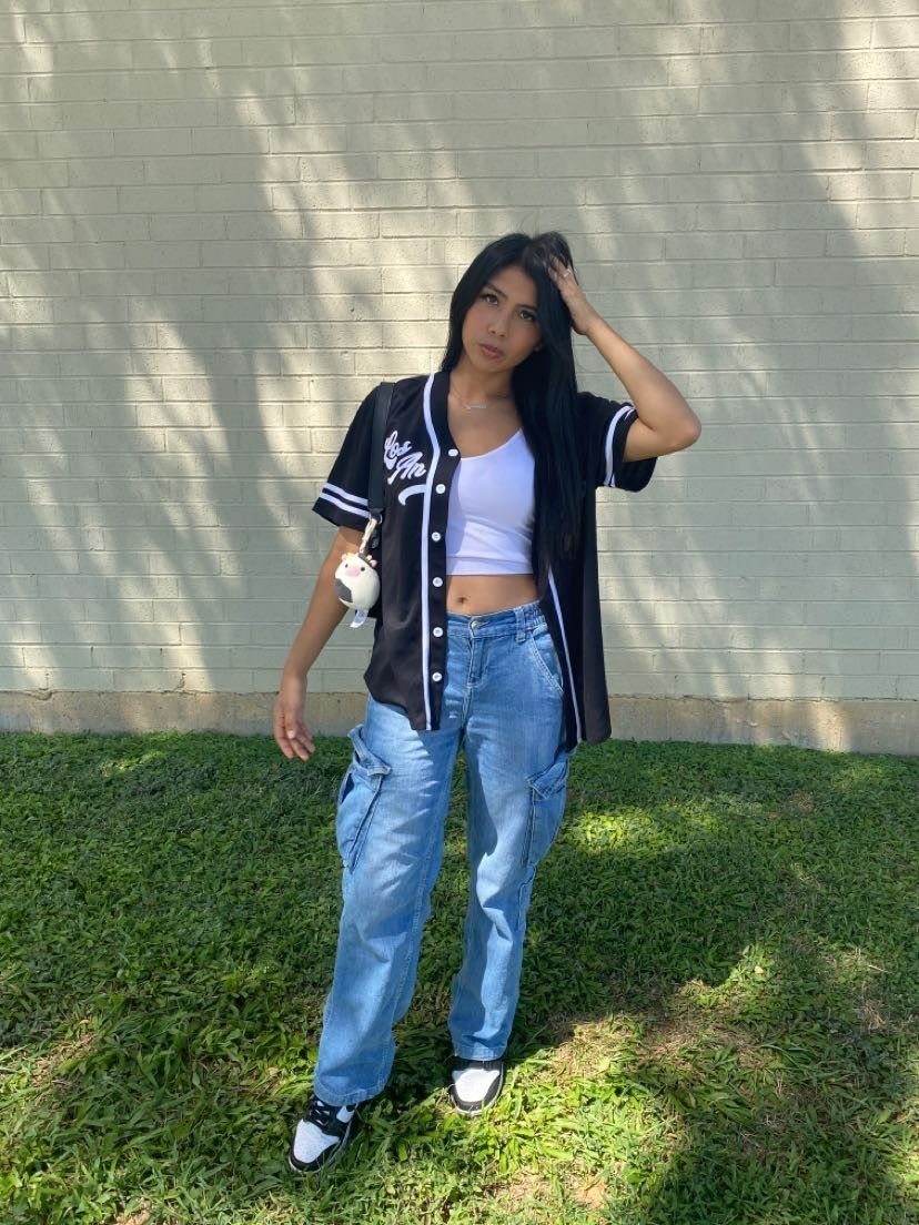 Woman wearing an open baseball jersey, white crop top, and blue denim cargo jeans standing outdoors.