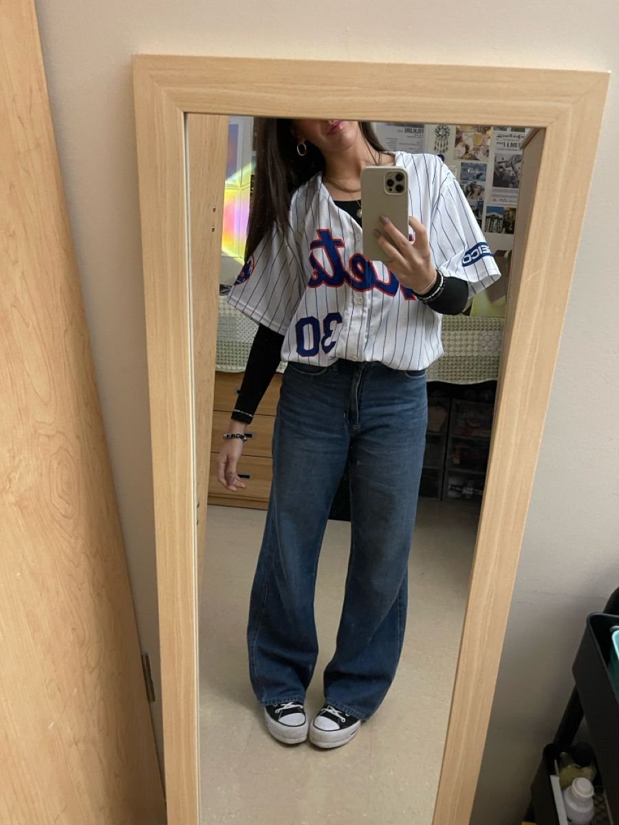 A woman posing in a mirror wearing a baseball jersey, blue jeans, and black sneakers.