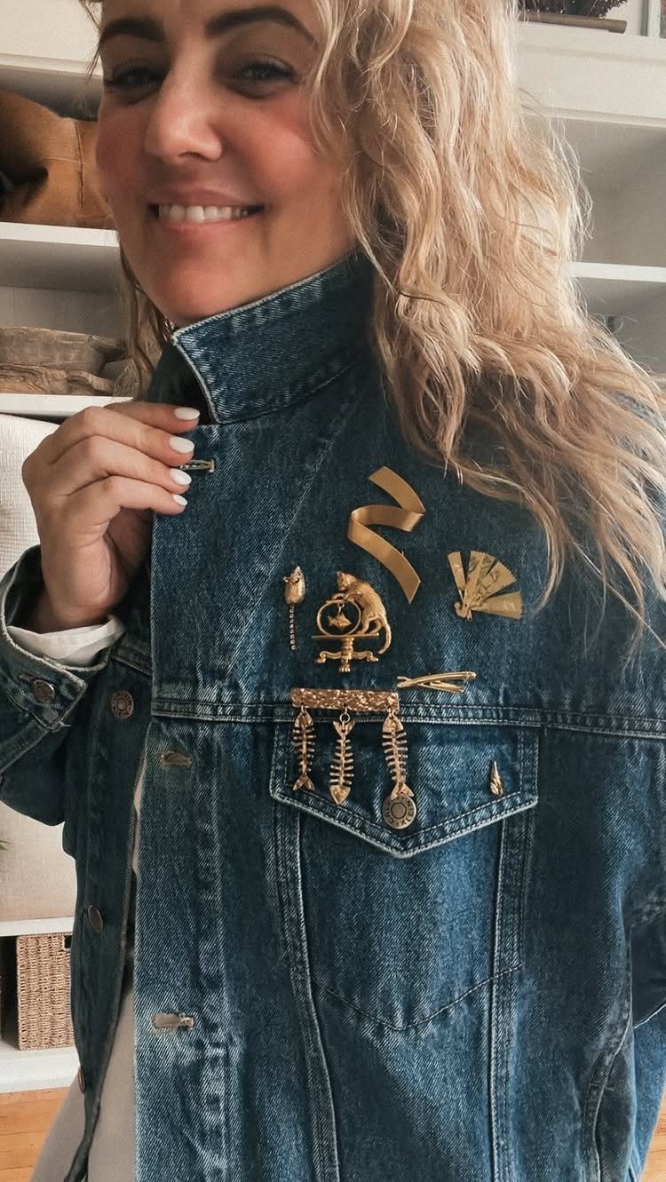A smiling blonde woman wearing a denim jacket decorated with various gold-toned decorative pins.