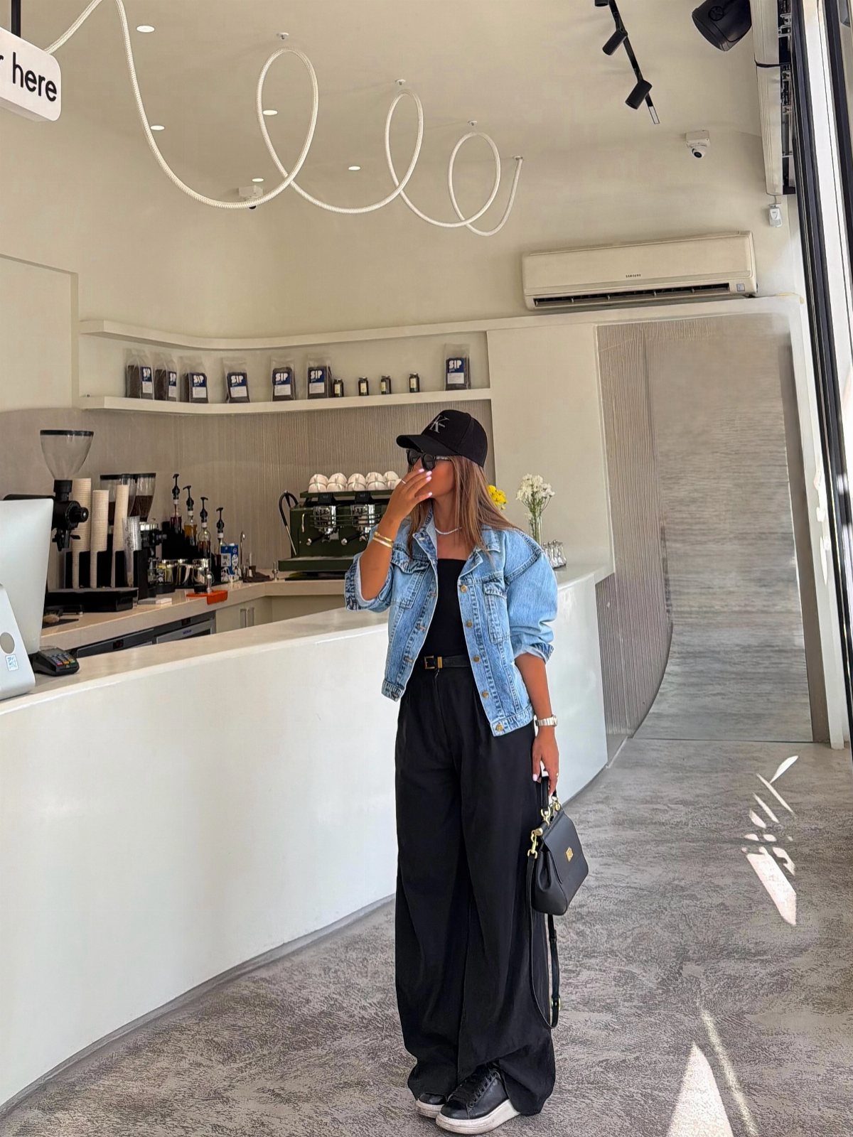 Woman wearing a light wash denim jacket, black top, and wide-leg trousers in a cafe.