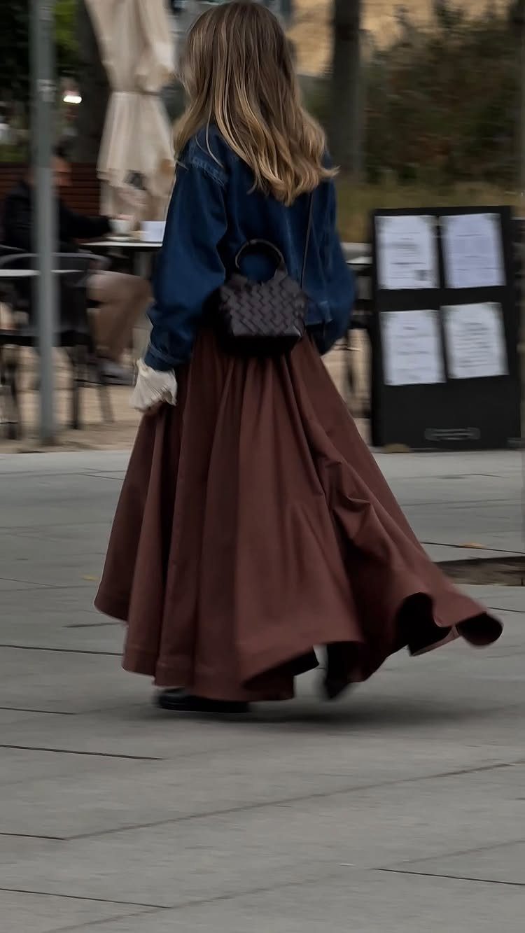 A woman walking in a blue denim jacket and a long brown flowing skirt.