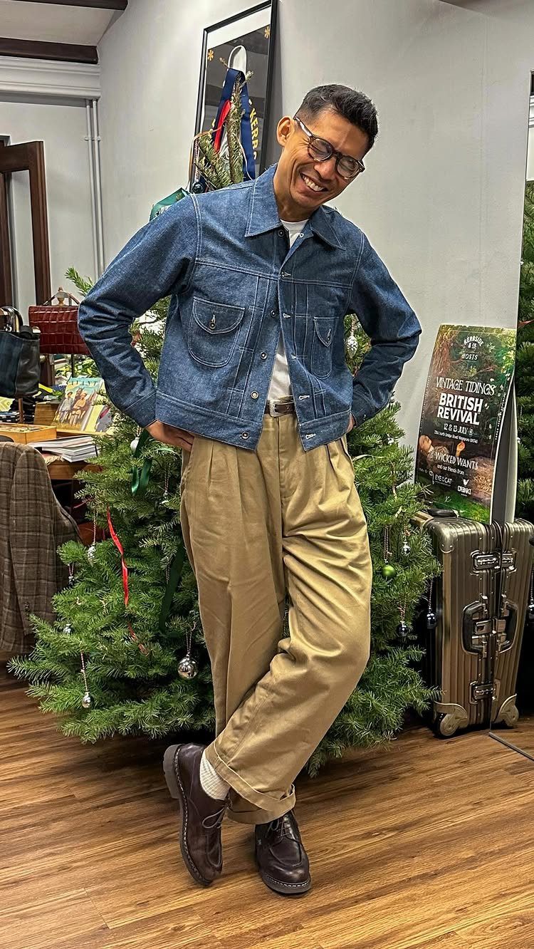 A man smiling in a blue denim jacket and tan pleated trousers.