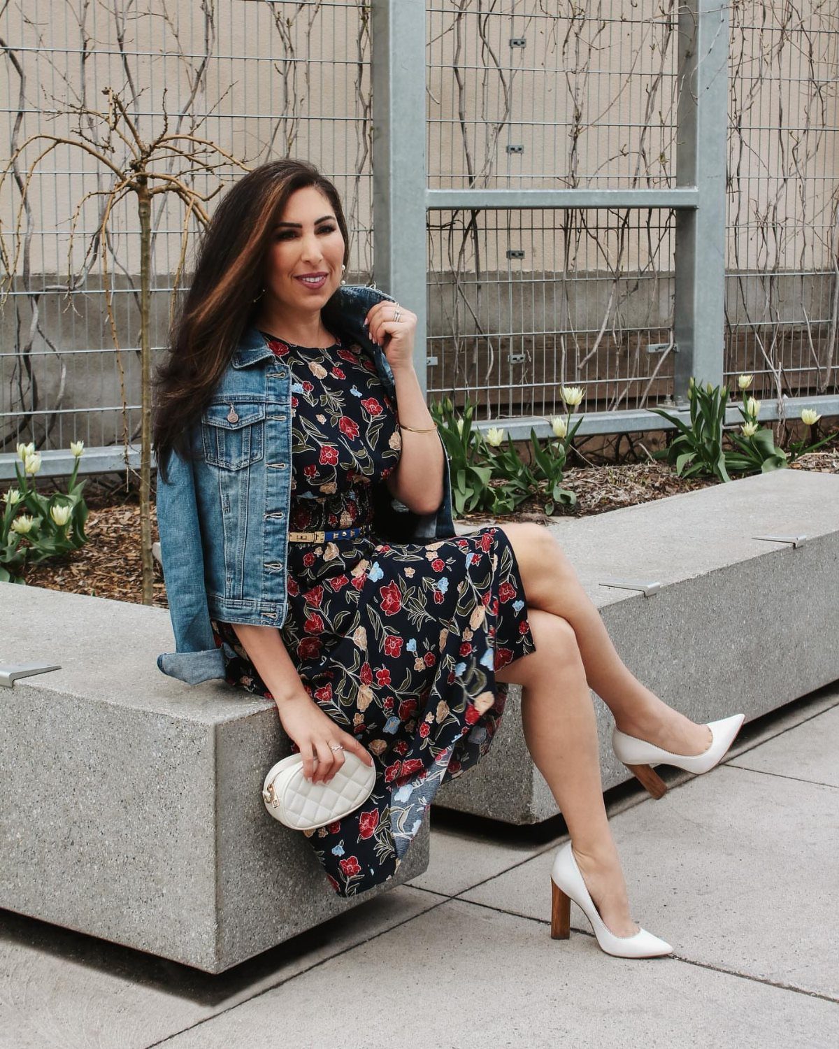 A woman sitting on a concrete bench wearing a floral dress and a denim jacket.