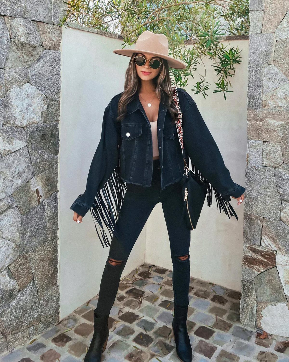 Woman posing in a black fringed denim jacket, black ripped jeans, and a tan wide-brimmed hat.