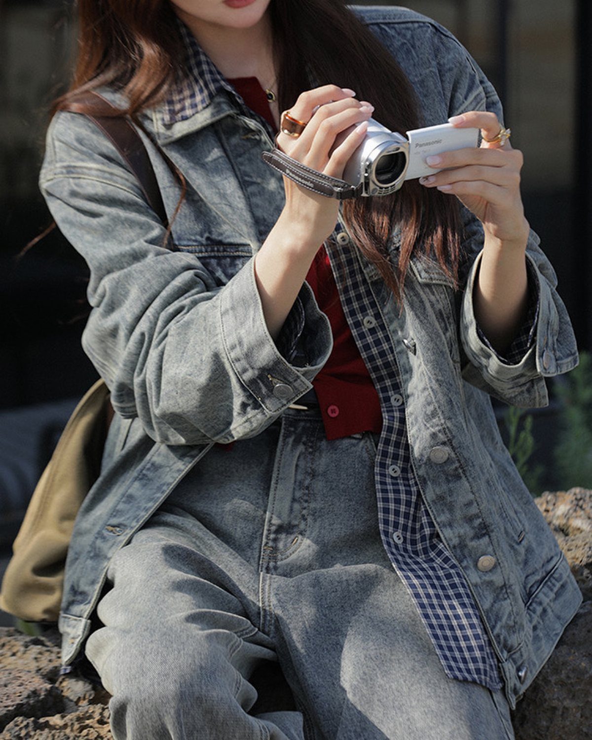 A person wearing a grey denim jacket and matching pants while holding a small camera.