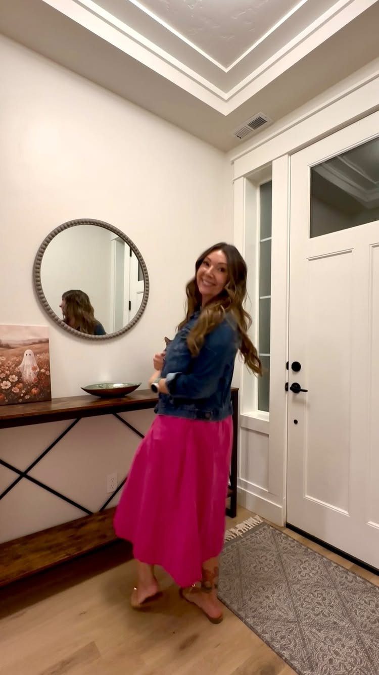 A smiling woman wearing a denim jacket over a bright pink midi dress in an entryway.