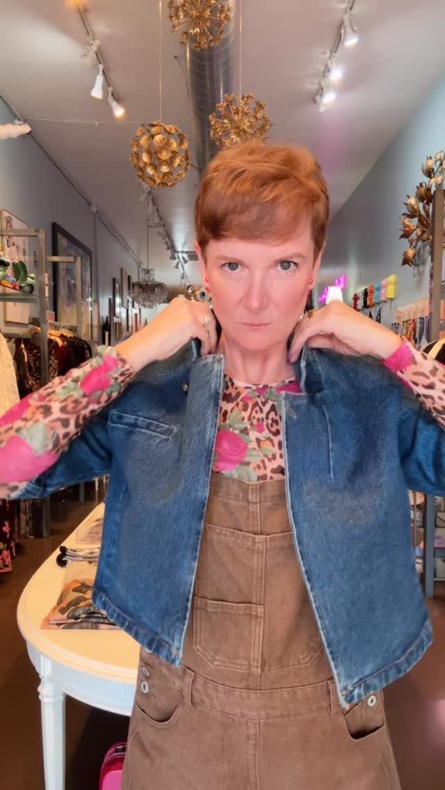 A woman wearing a blue denim jacket over floral print top and brown overalls.