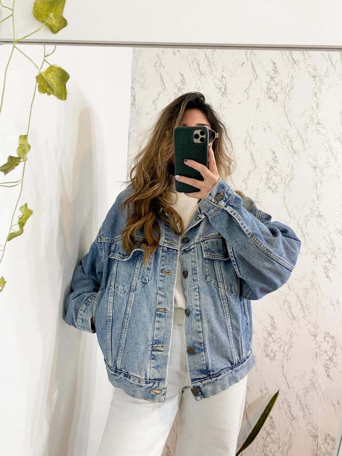 Woman in an oversized light wash denim jacket and white pants taking a mirror selfie.