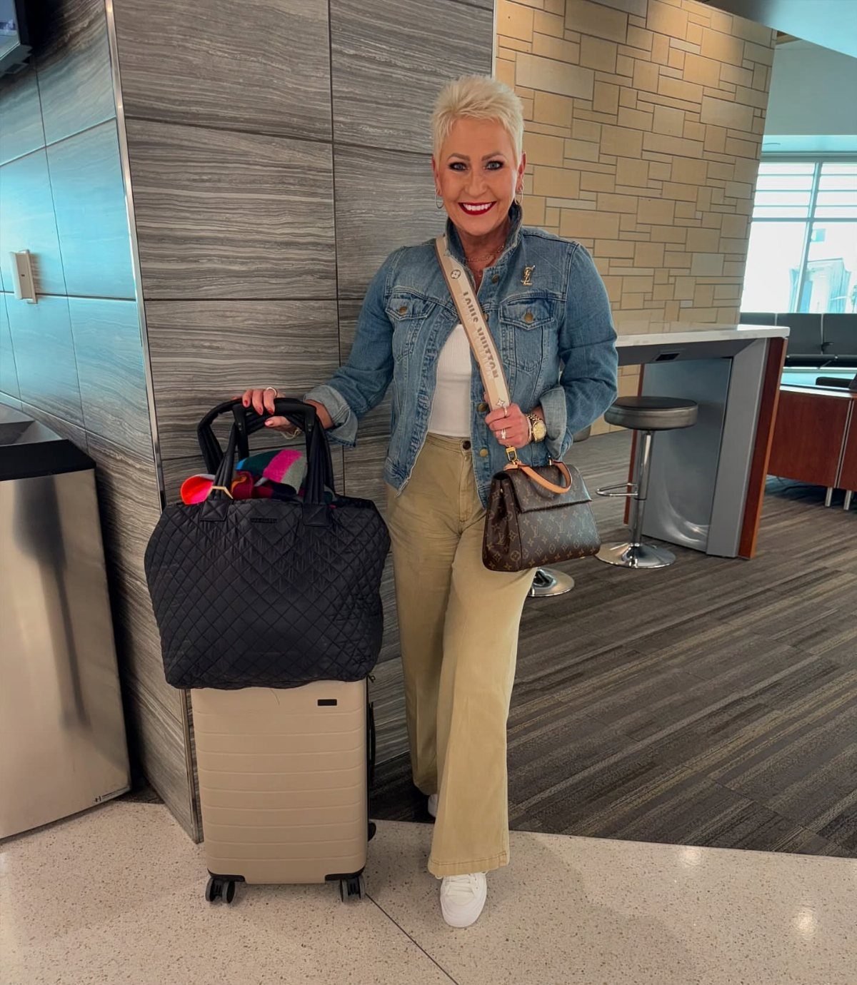 Smiling woman in a denim jacket standing with a suitcase and large black bag in an airport.
