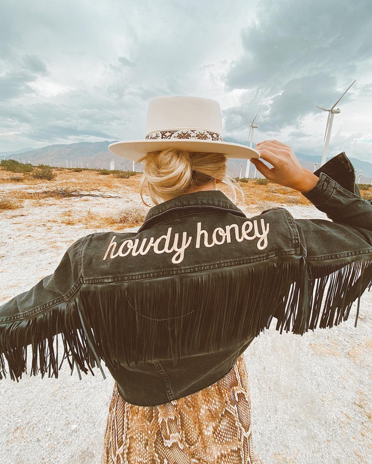 Woman in a fringed denim jacket and cowboy hat standing in a desert landscape.