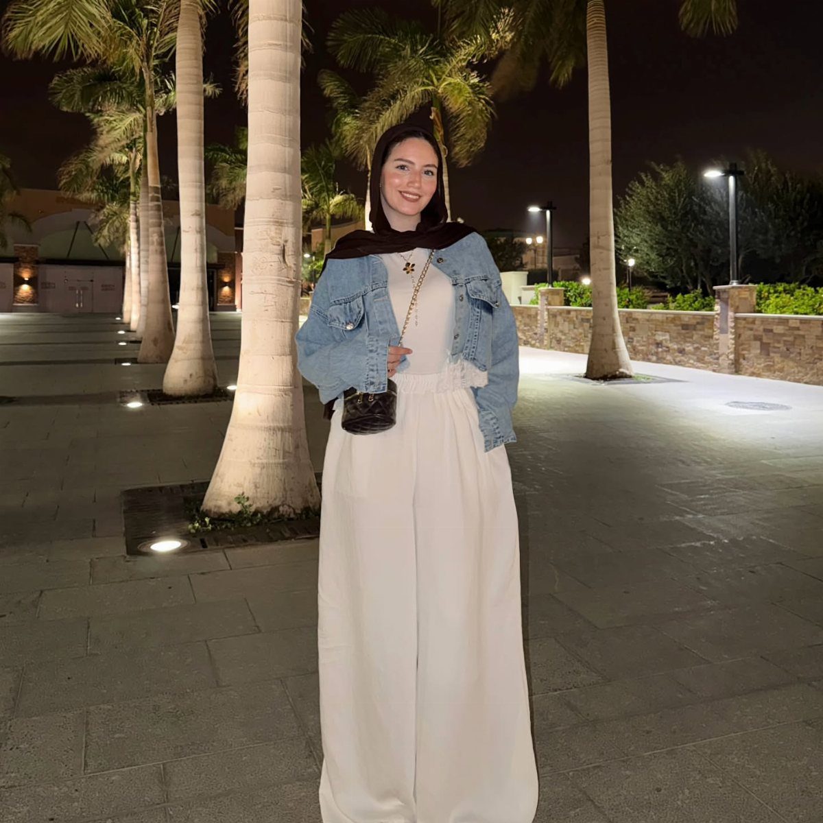 A woman wearing a denim jacket and white jumpsuit posing outdoors at night.
