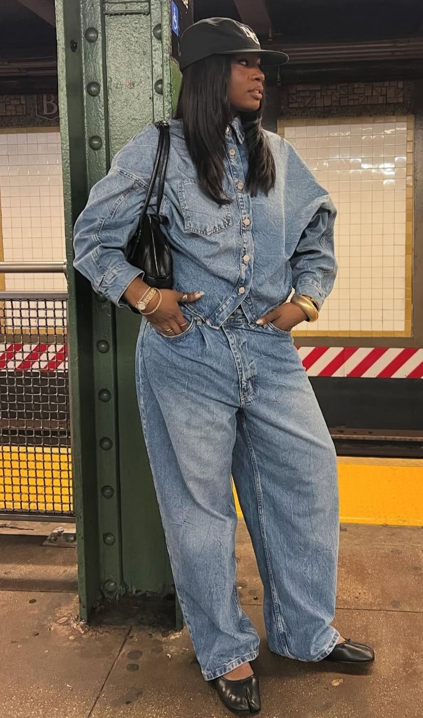 Woman in a denim jumpsuit and black square-toe flats standing in a subway station.