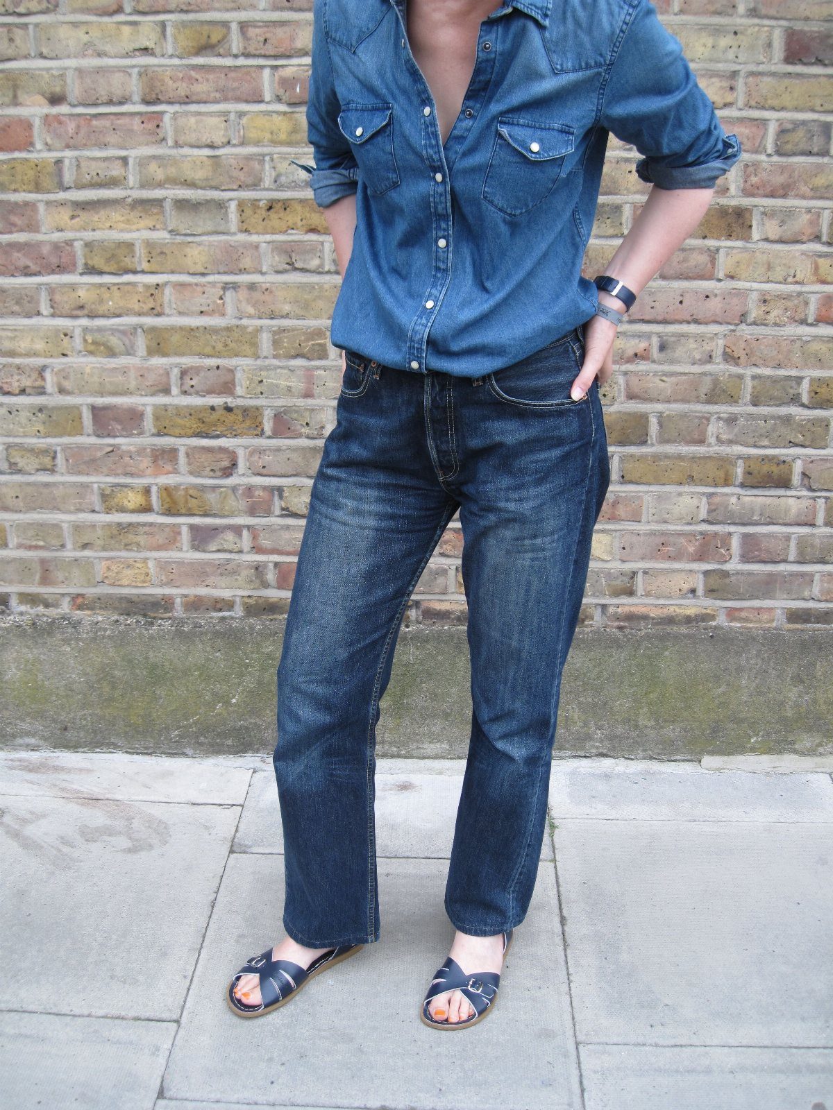Person wearing blue denim shirt, jeans, and navy blue flat sandals against a brick wall.