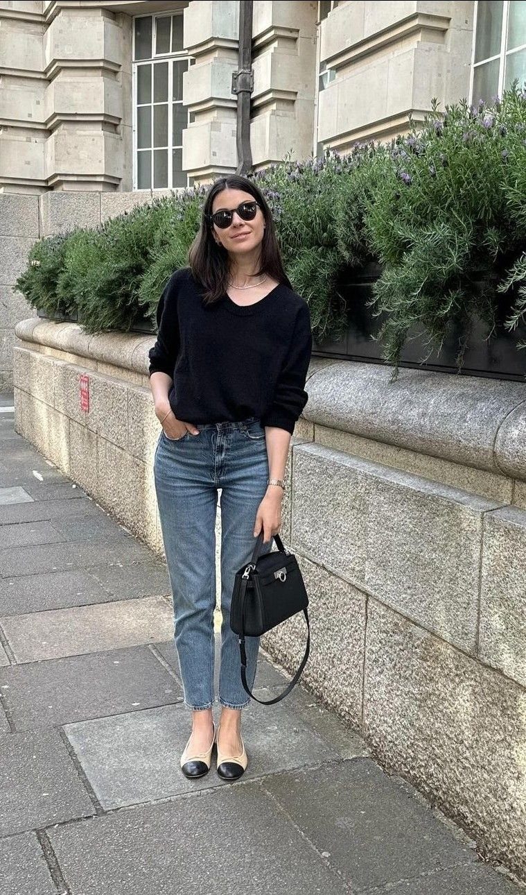 Woman wearing high-rise blue jeans, a black top, and black cap-toe ballet flats standing on a sidewalk.