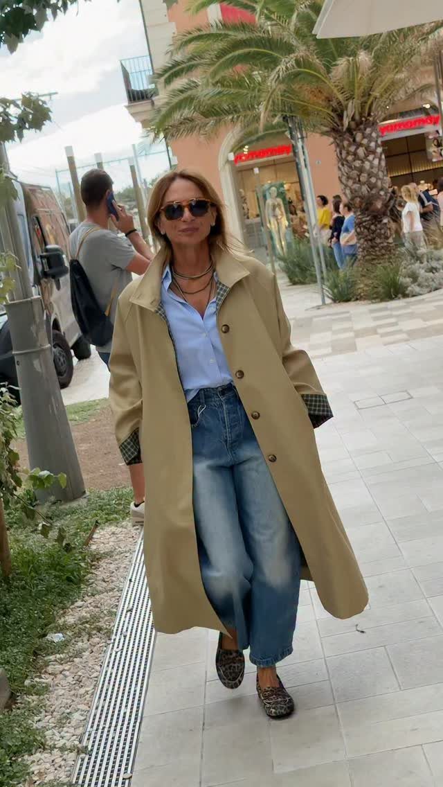 Woman in a tan trench coat, blue jeans, and leopard print loafers walking outdoors.