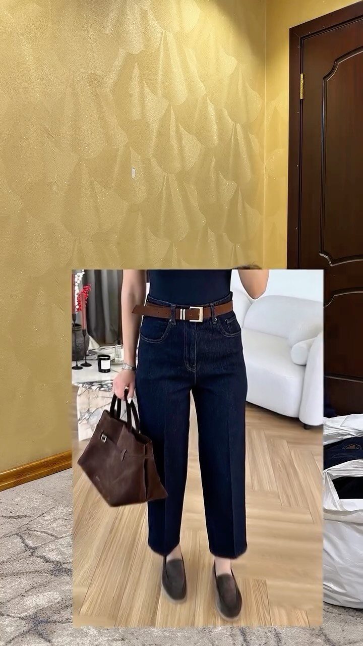 Person wearing dark blue high-waisted jeans, a black top, and a brown belt while holding a handbag.