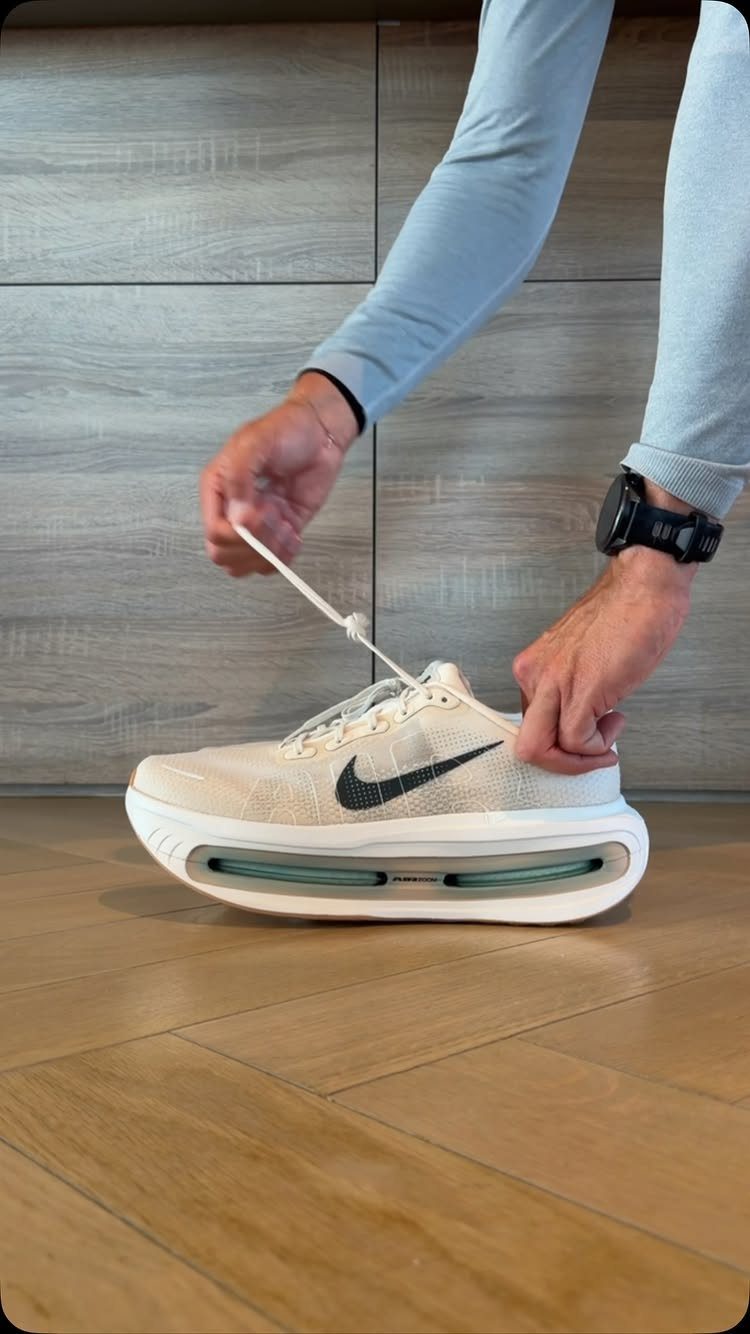 Hands tying the white laces of a cream Nike athletic shoe on a wooden floor.
