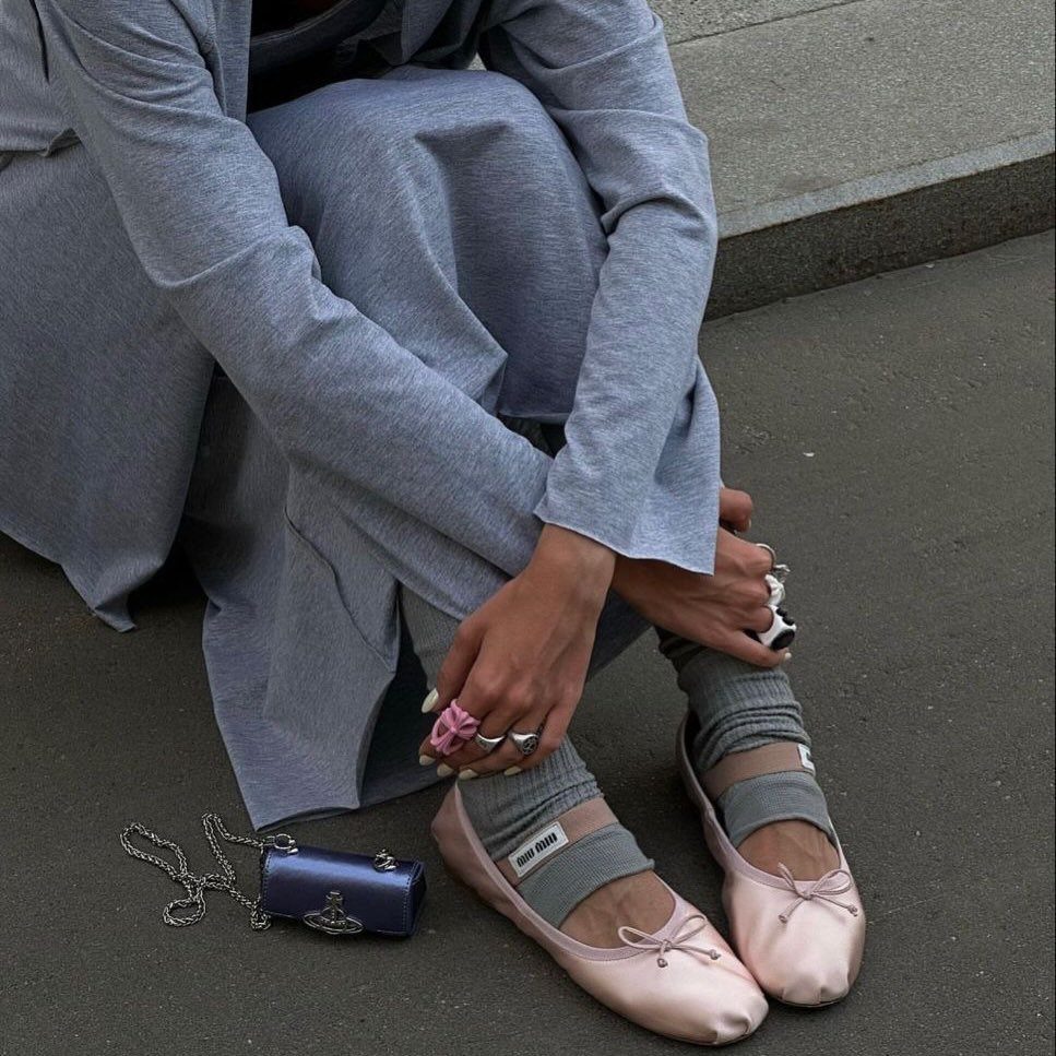 Person wearing pink satin ballet flats with bows and grey socks sitting on the ground.