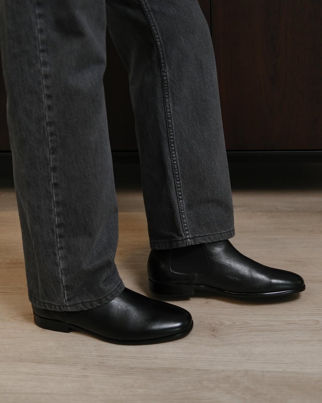 Black leather Chelsea boots worn with dark grey trousers on a wooden floor.
