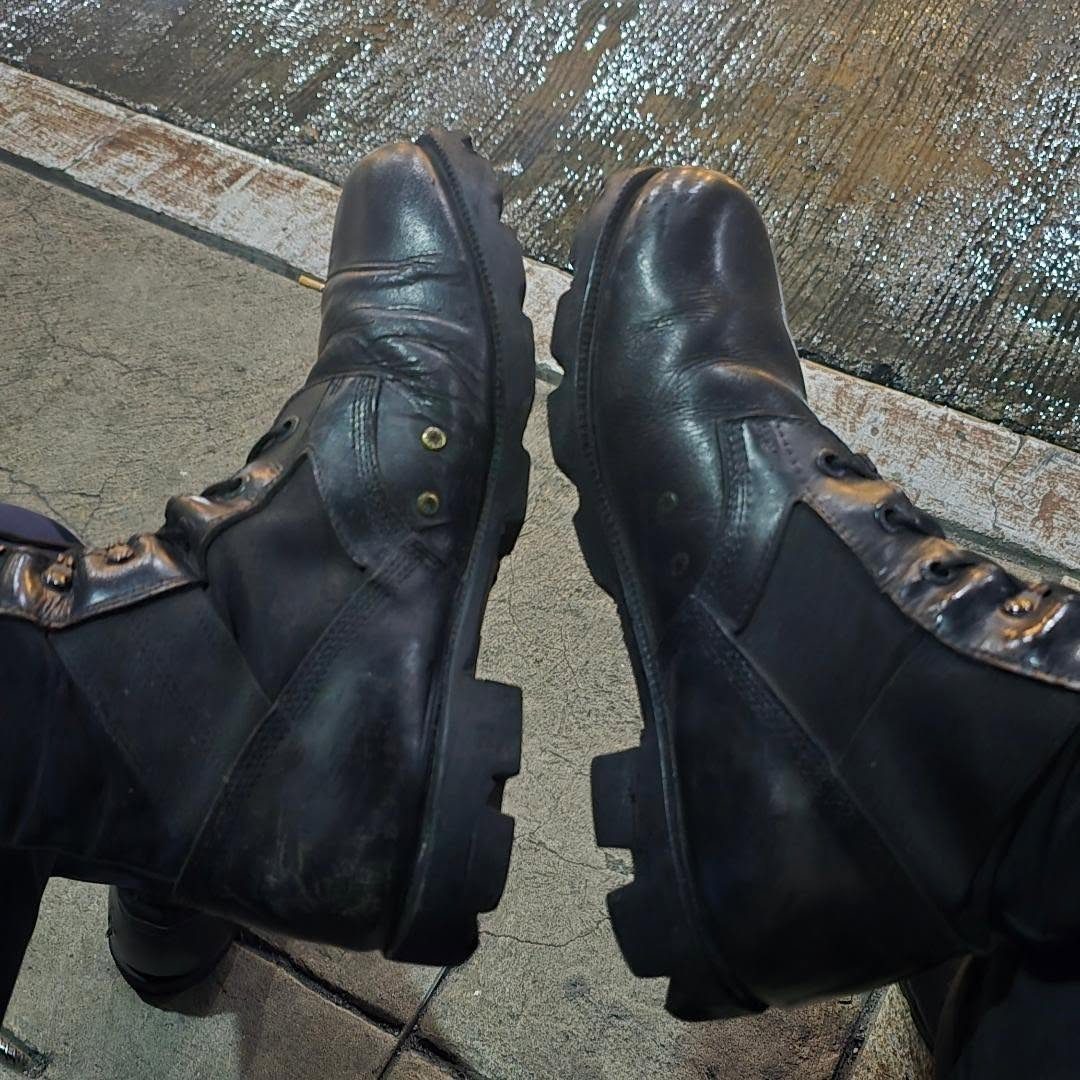 Black leather combat boots with thick lugged soles resting on a concrete sidewalk near a wet street.
