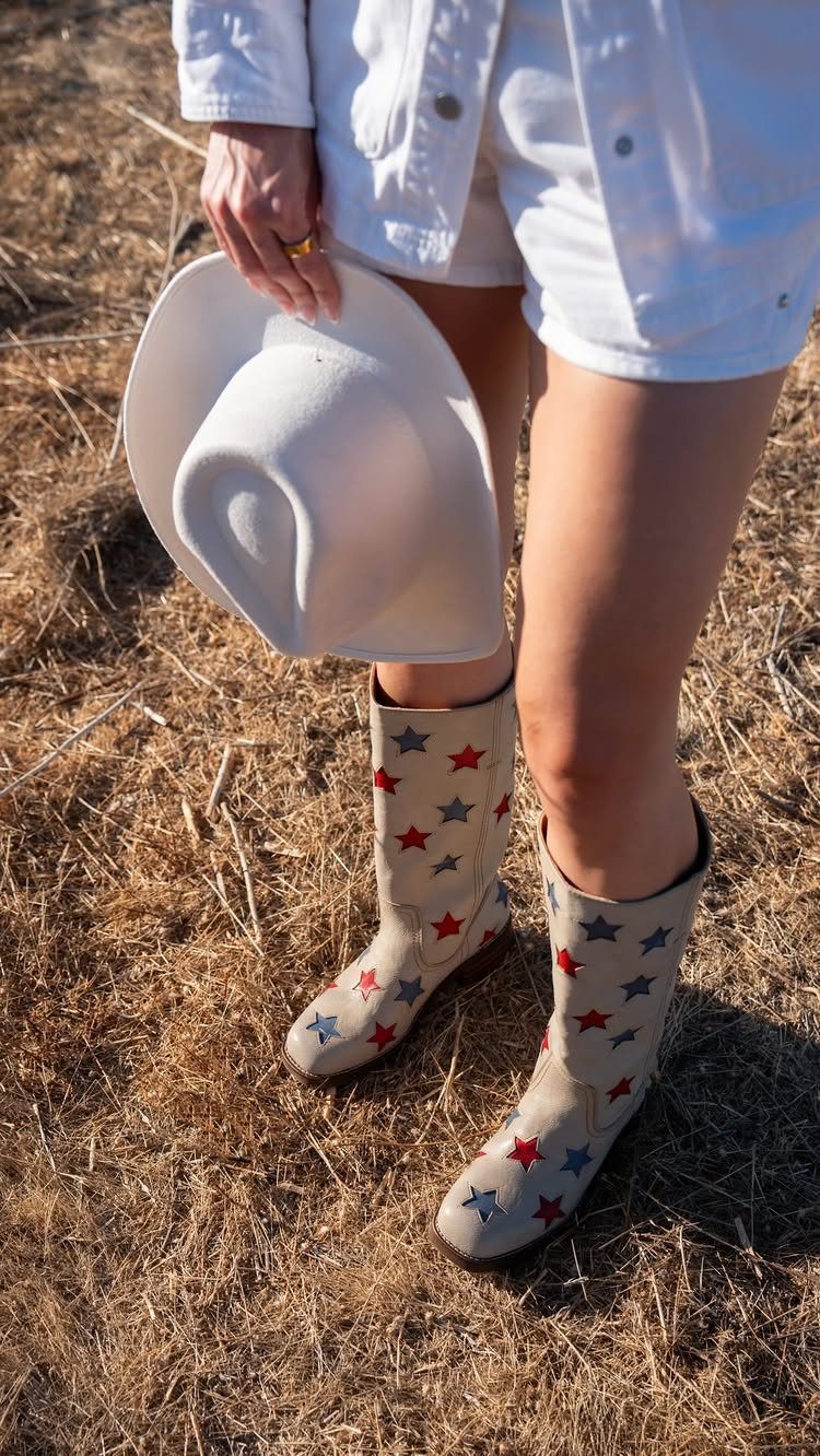 Person wearing cream cowboy boots with red and blue stars standing in dry grass.