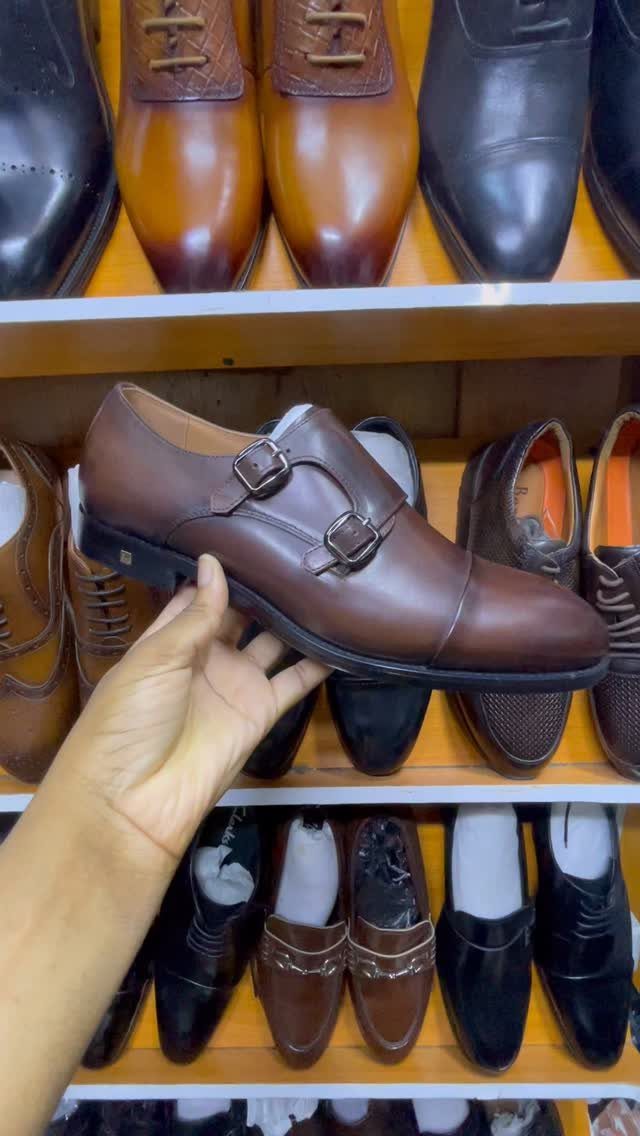 A hand holds a brown leather double monk strap dress shoe against a background of shoe shelves.