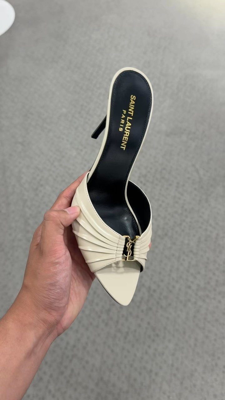 A hand holding a cream-colored pleated Saint Laurent mule with a gold logo.