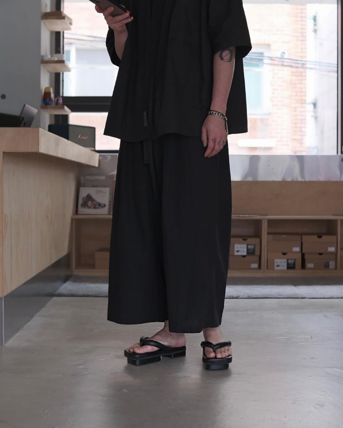 Person in black clothing wearing black platform thong sandals standing in a minimalist shop.