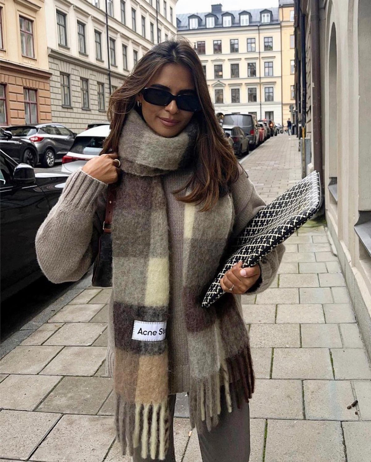 A woman wearing a large, thick plaid scarf and a beige sweater on a city street.