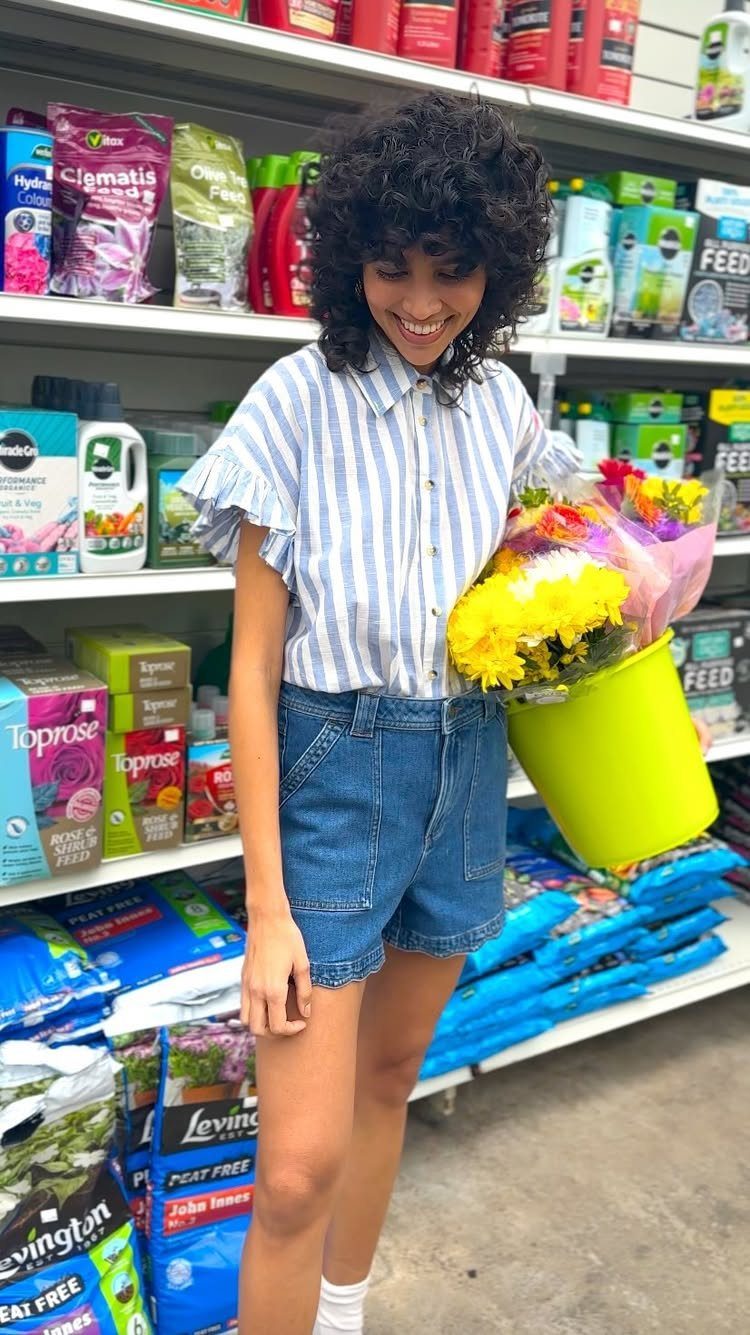 A smiling woman with curly hair wearing denim shorts and a striped shirt holding flowers.