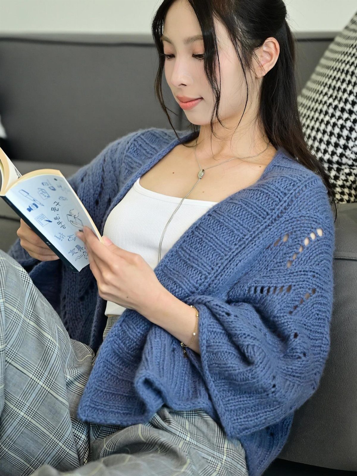A woman in a blue knit cardigan reads a book while sitting on a sofa.