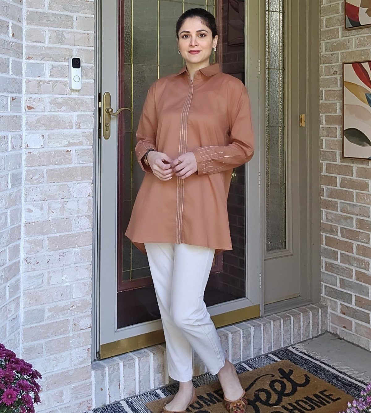 A woman posing in a terracotta tunic top and white pants by a doorway.