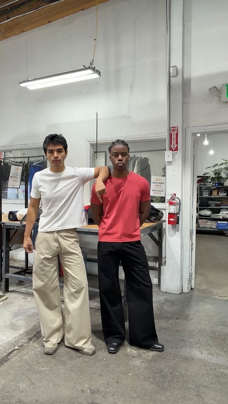 Two men posing in a workshop wearing t-shirts and wide-leg pants.