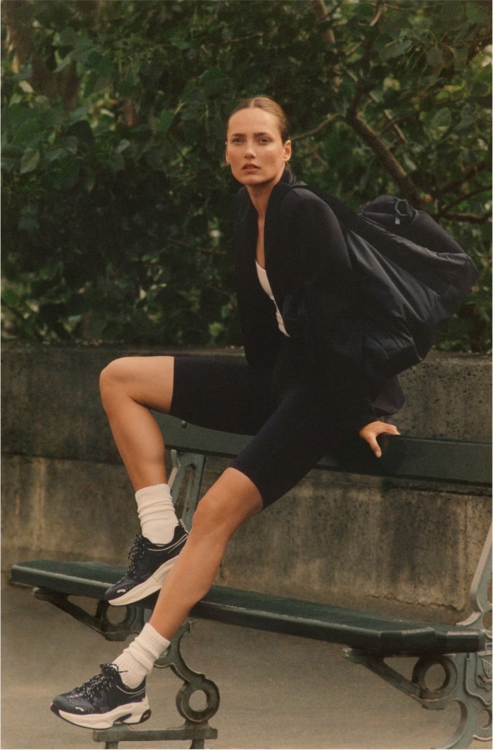 A woman in black athletic shorts and sneakers sits on a park bench with a large black bag.