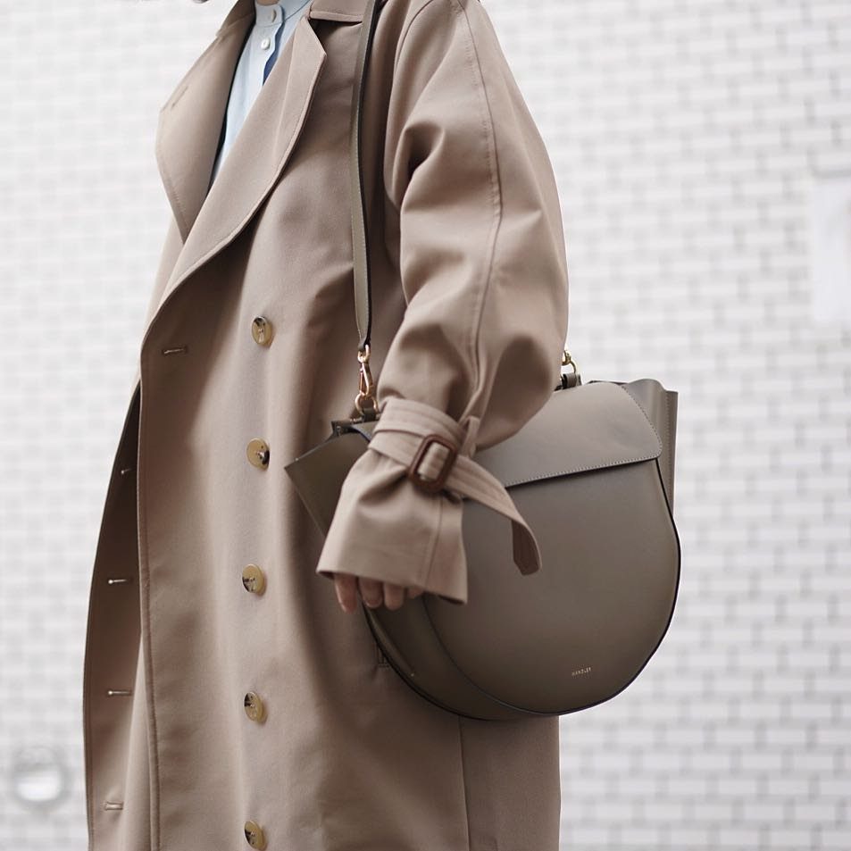 A person wearing a beige trench coat and carrying a large taupe leather bag.