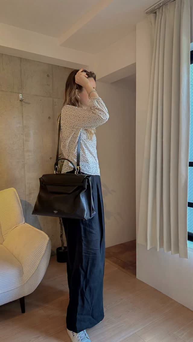 Woman in patterned blouse and wide black trousers carrying a large black leather handbag.