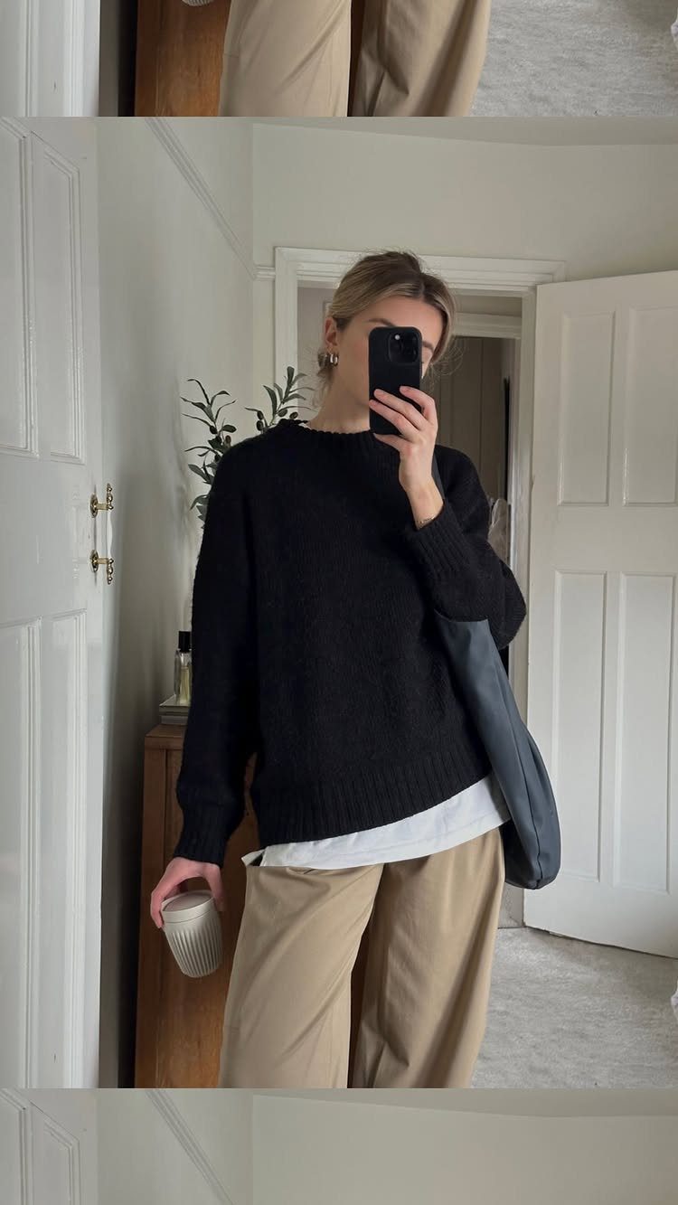 Woman in black sweater, white shirt, and tan trousers posing for a mirror selfie.